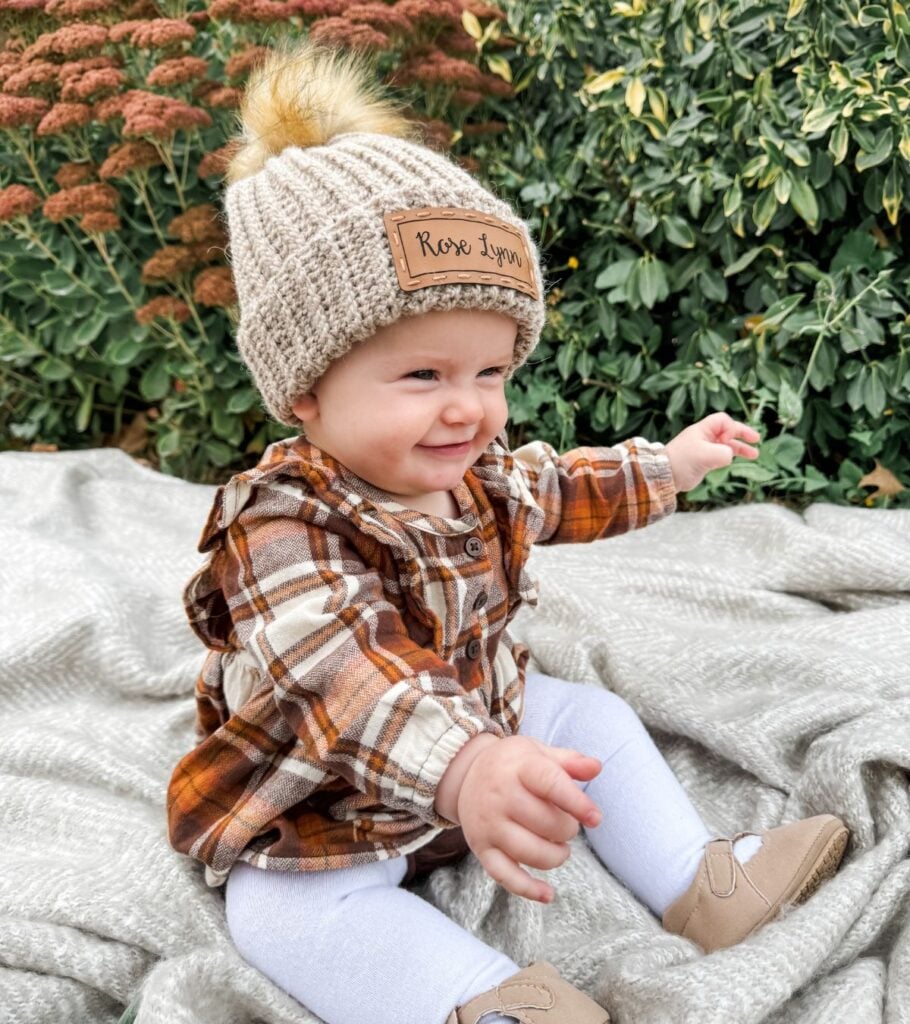 A baby in a plaid hat sitting on a blanket, holding a crochet bag.