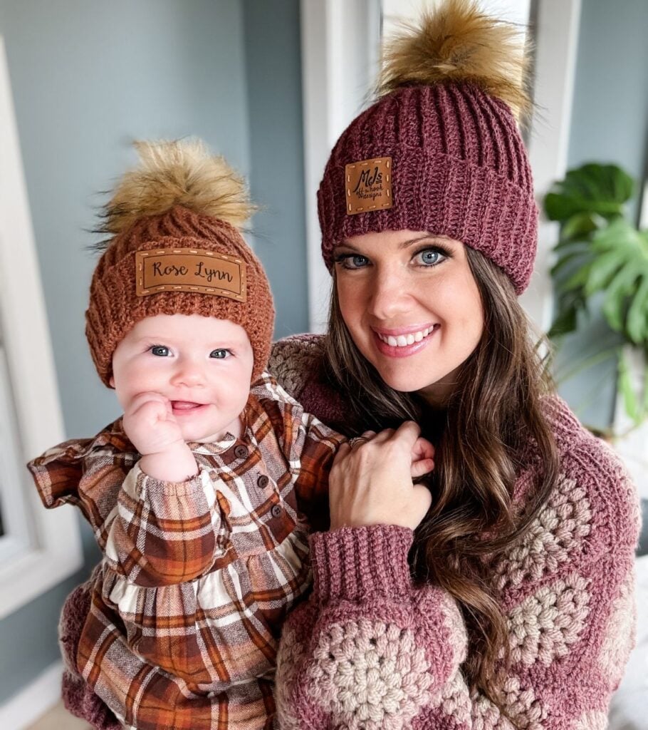 A woman holding a baby wearing a burgundy pom pom beanie and carrying a crochet bag.