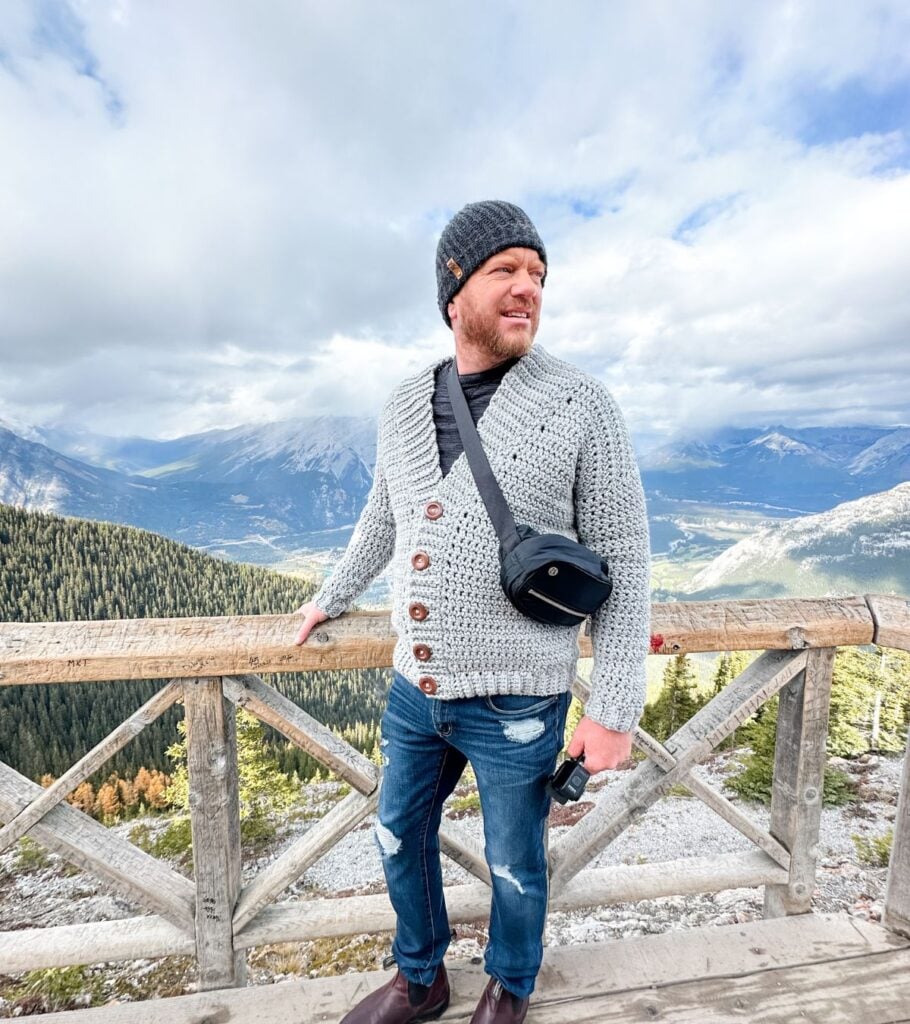 A Rocky Ridge Crochet Hat customer is captured standing on a wooden bridge with mountains in the background.