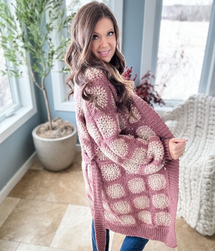 A woman with long brown hair wearing a pink and beige crochet coatigan poses indoors near large windows and a potted plant.