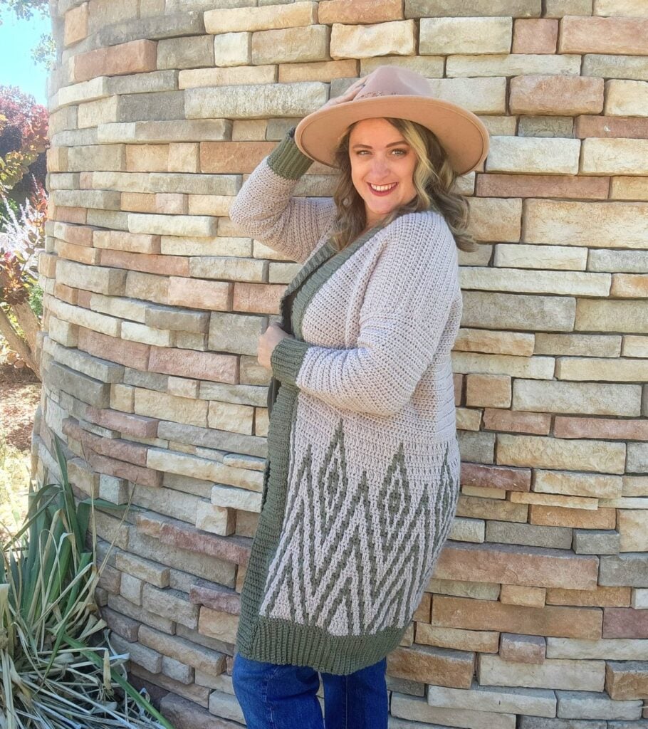 A woman wearing a hat and cardigan standing next to a brick wall.