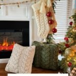 A fireplace decorated for Christmas with stockings, Christmas crochet ornaments, two cabled crochet pillows in cream and green, and a lit tree beside a sign reading "Cozy Cottage Cabled Pillow.