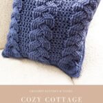 A blue crocheted pillow with a cabled pattern rests on a beige fabric surface, adding charm to your space. Text below reads: "Cozy Cottage Cabled Pillow, mjsoffthehookdesigns.com." Pair with a Christmas Crochet Ornament for festive flair.