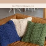 Three knit pillows in blue, cream, and green with a cabled pattern are arranged on a brown leather couch, offering a cozy cottage vibe. Perfect complement to your Christmas crochet ornament décor. Text above reads "Cozy Cottage Cabled Pillow.