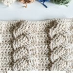 A textured crochet pillow with cable patterns is shown, with four skeins of yarn in cream, brown, blue, and green above it. Text reads: Cozy Cottage Cabled Pillow—a perfect match for your next Christmas Crochet Ornament project.