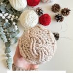 A hand holds a beige crocheted Christmas Crochet Ornament bauble, with red and white yarn balls, pinecones, and greenery in the background. Text reads: "Cozy Cottage Cabled Baubles Crochet Pattern & Video.