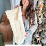 Woman holding a large cream-colored cabled stocking in front of a decorated Christmas tree. Text at the bottom reads "Cozy Cottage Cabled Stocking Crochet Pattern & Video.