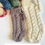 Two crocheted cabled stockings, one cream and one taupe, with name tags, displayed beside yarn balls, a candle, and crochet hooks—perfect for showcasing your favorite cabled stocking style this holiday season.