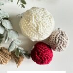 Three Christmas Crochet Ornaments—cabled baubles in ivory, red, and taupe—are displayed on a white surface, accented by green foliage and twine nearby.