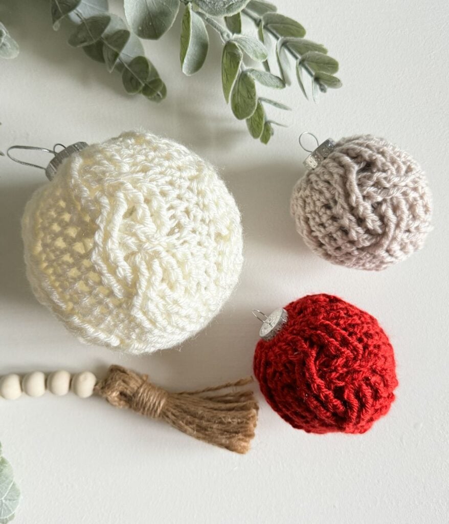 Three Christmas crochet ornaments in white, beige, and red are arranged on a white surface near a beaded jute tassel and green foliage.