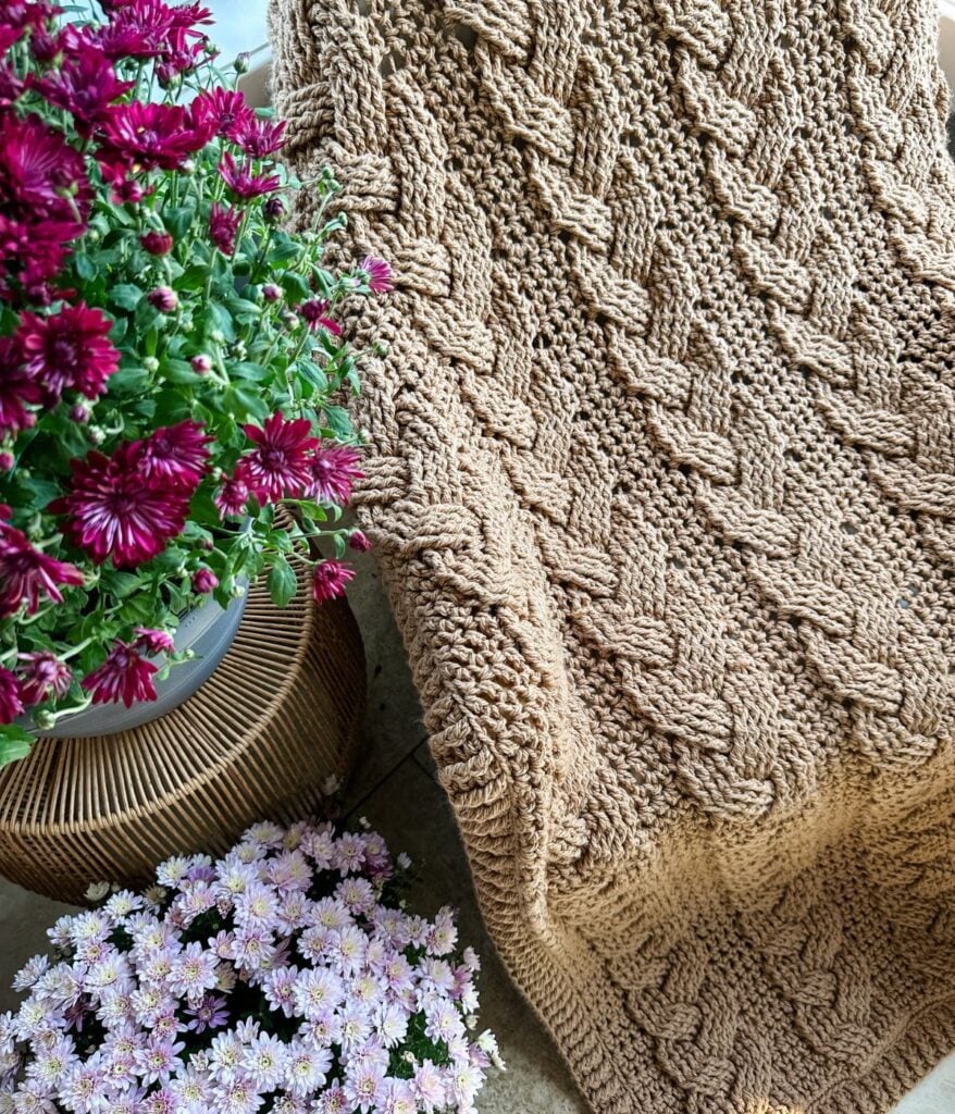 A cabled throw blanket in brown is draped over a chair, showcasing its intricate knit beside pots of purple and light pink chrysanthemums.