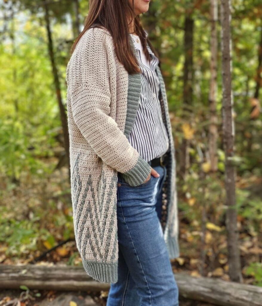 A person stands outdoors wearing a beige and green mosaic crochet cardigan over a striped shirt and blue jeans, surrounded by trees and foliage.