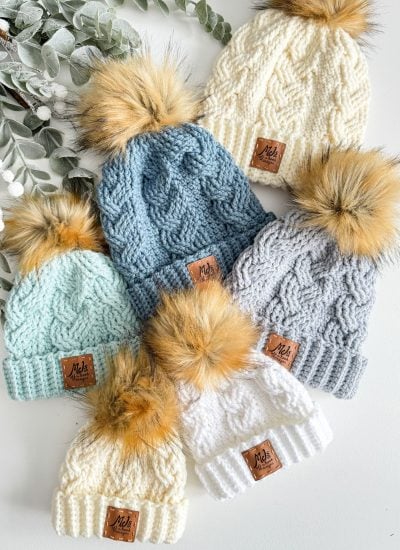 Six cabled hats with faux fur pom-poms in cream, blue, gray, and mint are arranged on a white surface next to greenery.
