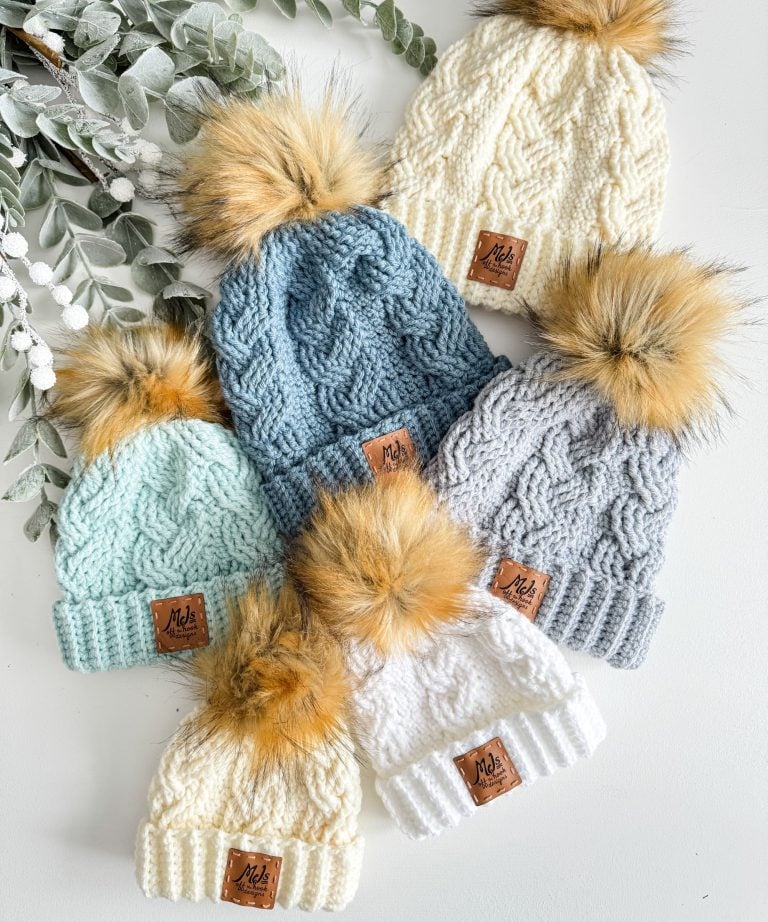 Six cabled hats with faux fur pom-poms in cream, blue, gray, and mint are arranged on a white surface next to greenery.