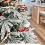 A snowy artificial Christmas tree decorated with granny square ornaments, crochet baubles, and wooden beads stands in a bright, modern kitchen. Text reads "Holiday Weave Baubles Crochet Pattern & Video.