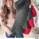 A woman holds up large crocheted holiday stockings in green, red, and white, adorned with tassels and a granny square ornament. Text below reads: "Holiday Weave Stocking Crochet Pattern & Video.”.