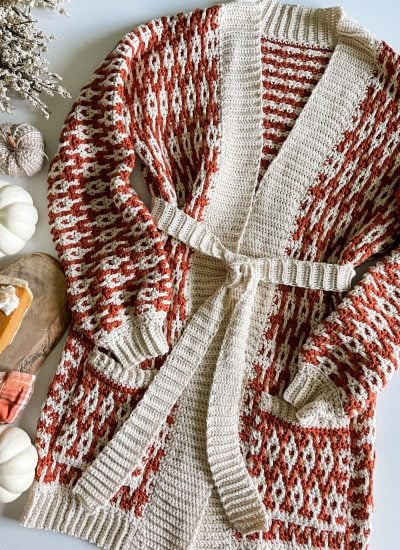 A knitted cardigan with a red and white geometric pattern and a belt is laid out on a table beside a cozy cabled hat, white pumpkins, dried flowers, and a slice of pumpkin pie on a wooden board.