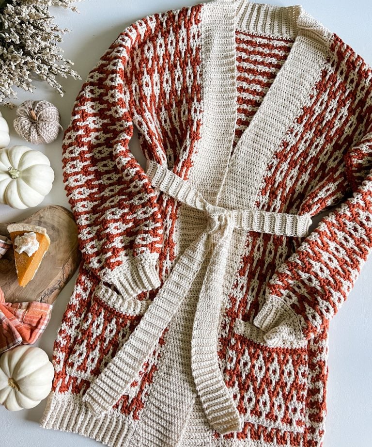A knitted cardigan with a red and white geometric pattern and a belt is laid out on a table beside a cozy cabled hat, white pumpkins, dried flowers, and a slice of pumpkin pie on a wooden board.