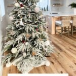 A decorated Christmas tree with ornaments, including a charming Granny square ornament, and a woven tree skirt stands in a modern, bright kitchen with wooden floors and bar stools.