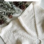 A white woven holiday stocking with a brown ribbon, surrounded by pinecones, greenery, and a charming granny square ornament, displayed next to text reading "Holiday Weave Stocking Crochet Pattern & Video.