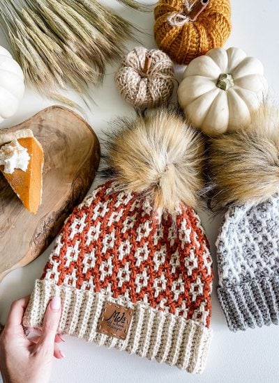 Two cabled hats with faux fur pom-poms lie on a table with decorative pumpkins, wheat stalks, and a slice of pumpkin pie on a wooden tray.