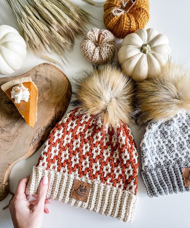Two cabled hats with faux fur pom-poms lie on a table with decorative pumpkins, wheat stalks, and a slice of pumpkin pie on a wooden tray.