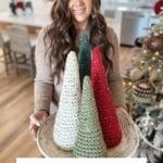 A woman holds a tray with four crochet holiday trees in white, green, and red. Text reads: "Rustic Holiday Tree: Crochet Pattern & Video." A decorated Christmas tree and a granny square ornament are in the background.