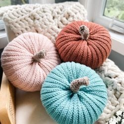 Three knitted pumpkins in pink, orange, and teal are arranged on a chair with a chunky knit blanket and a cozy cabled hat near a window.