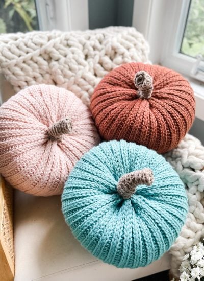 Three knitted pumpkins in pink, orange, and teal are arranged on a chair with a chunky knit blanket and a cozy cabled hat near a window.