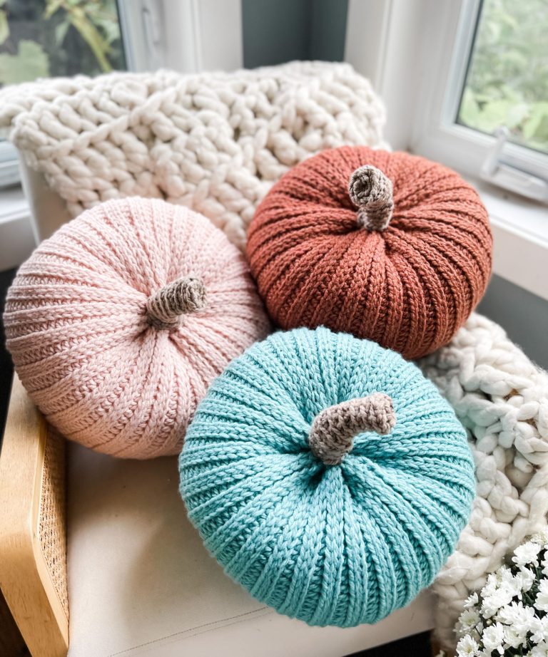 Three knitted pumpkins in pink, orange, and teal are arranged on a chair with a chunky knit blanket and a cozy cabled hat near a window.