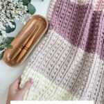 A hand holds a knitted blanket with cream, pink, and mauve stripes next to crochet hooks, a wooden tray, and a charming granny square ornament, surrounded by greenery. Text reads: "Snuggle Season Blanket Crochet Pattern & Video.