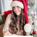 Woman wearing a red and white crocheted Santa hat poses indoors in front of a decorated Christmas tree with a granny square ornament. Text reads: “SANTA HAT Crochet Pattern & Video.”.