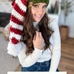 A woman wearing a red, white, and green crocheted elf hat with a white pompom sits indoors, smiling at the camera. Text reads: "Elf Hat Crochet Pattern & Video." Perfect to pair with a granny square ornament for festive flair!.