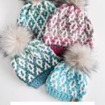 Four crocheted beanies with pom-poms, each featuring a mosaic pattern in blue, teal, or pink and white, are arranged on a white surface like a cozy display of granny square ornaments.