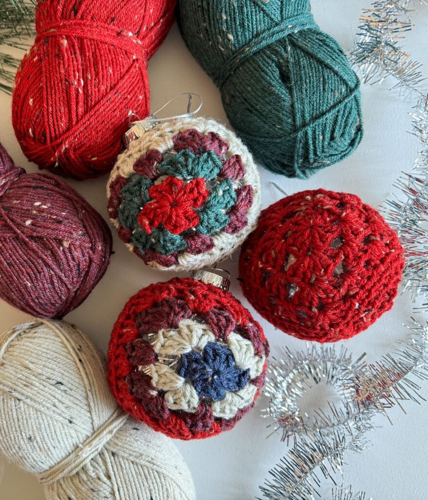 Four skeins of yarn in red, green, maroon, and cream are arranged with three crocheted ornaments—including a charming granny square ornament—and silver tinsel on a light surface.
