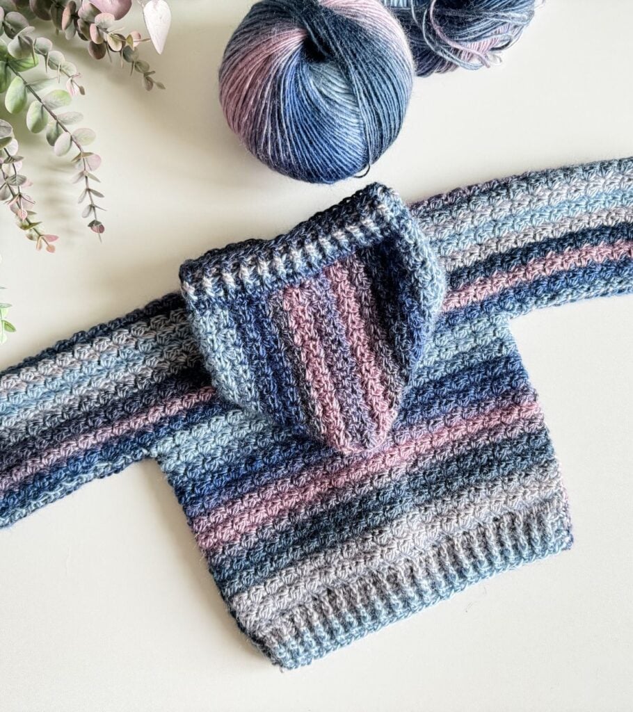 A crocheted cardigan with blue and pink yarn next to a flower.