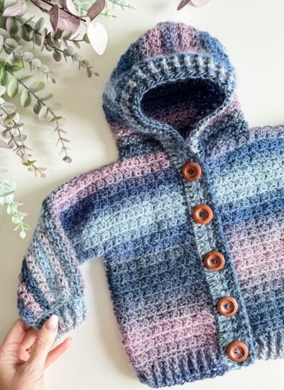 A hand holding a crocheted Granny Stitch hooded jacket.