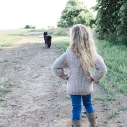 A little girl wearing a Granny Stitch Scarf standing on a dirt road looking at a dog.