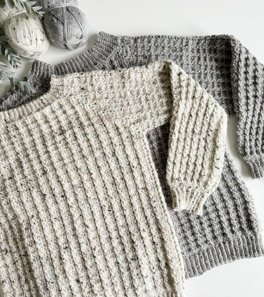 Two knitted sweaters on a table, one next to yarn.