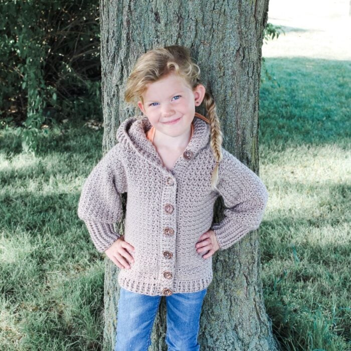 A little girl is standing next to a tree wearing a knitted cardigan and a Granny Stitch Scarf.