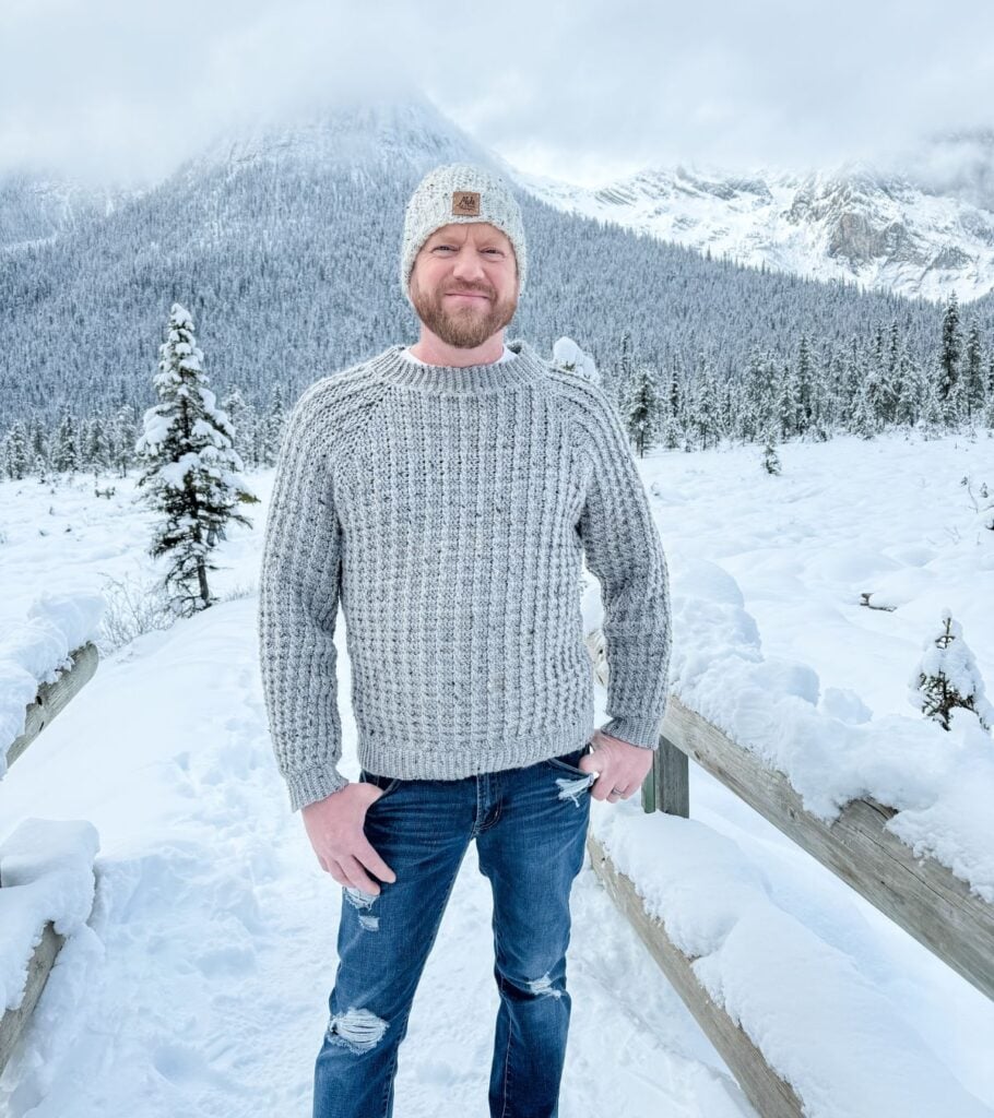 A man standing in the snow wearing a crochet hat, sweater, and jeans.