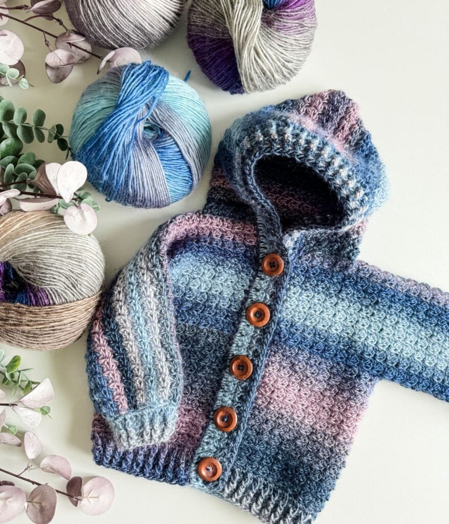 A Children's Hooded Cardigan with wooden buttons is displayed beside three balls of multicolored yarn and some eucalyptus branches on a white surface.