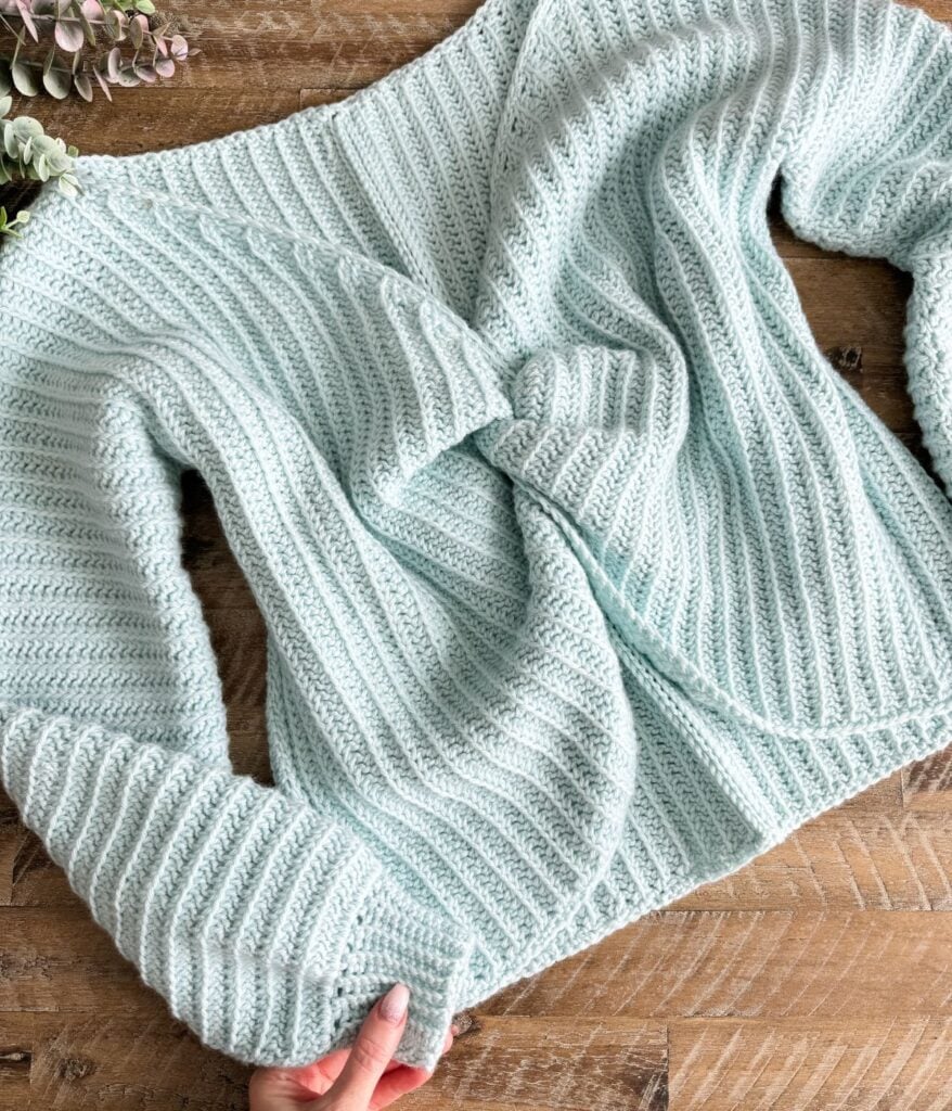 A light blue ribbed knit sweater, styled like the Sow Pretty Twist Top, is laid out on a wooden surface, with a hand holding one sleeve in the lower corner.