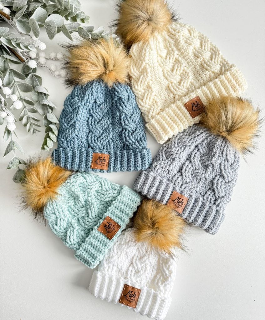 Five cozy crochet hats in blue, cream, gray, mint, and white with faux fur pom-poms are arranged on a white surface next to greenery.