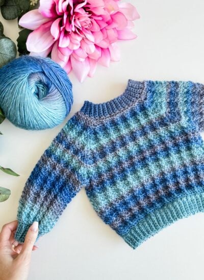 A men's pullover sweater with blue and grey hues, adorned with yarn and floral accents.