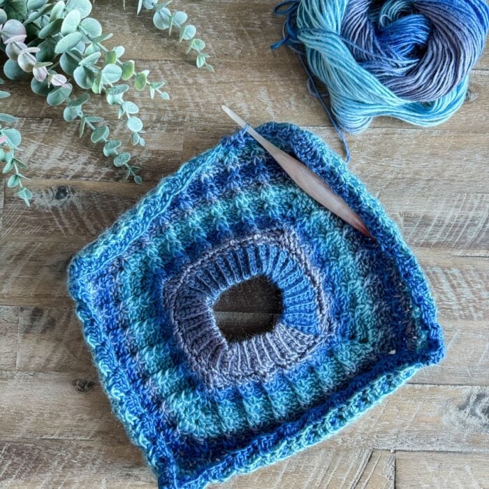A blue crocheted square on a wooden table, suitable for a men's pullover.