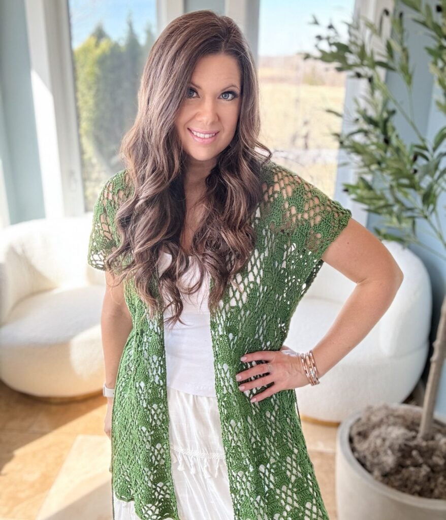 A woman with long wavy hair, wearing a green crochet shawl and white top, smiles in a bright, sunlit room with a plant and white furniture in the background.