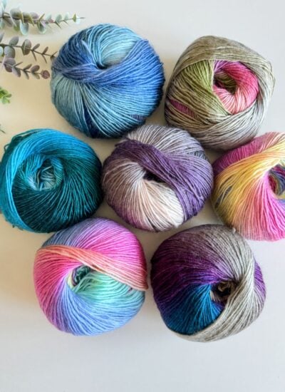 Assorted colorful yarn skeins arranged in a pattern on a light surface with greenery to the side, ready for a crochet cardigan project.