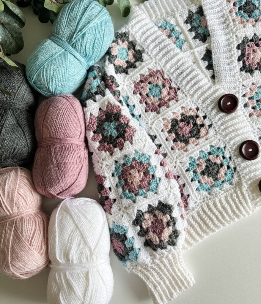 Colorful yarn balls next to a crocheted granny square blanket with floral patterns, displayed on a white surface.