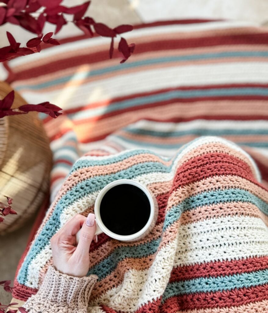 A person wrapped in a striped blanket, holding a cup of coffee, showcases the easiest children's crochet pullover.